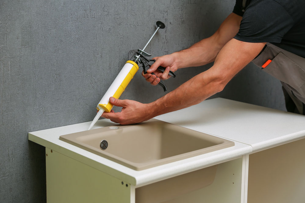 A man uses a sealant applicator gun to seal around the edge of a sink basin.